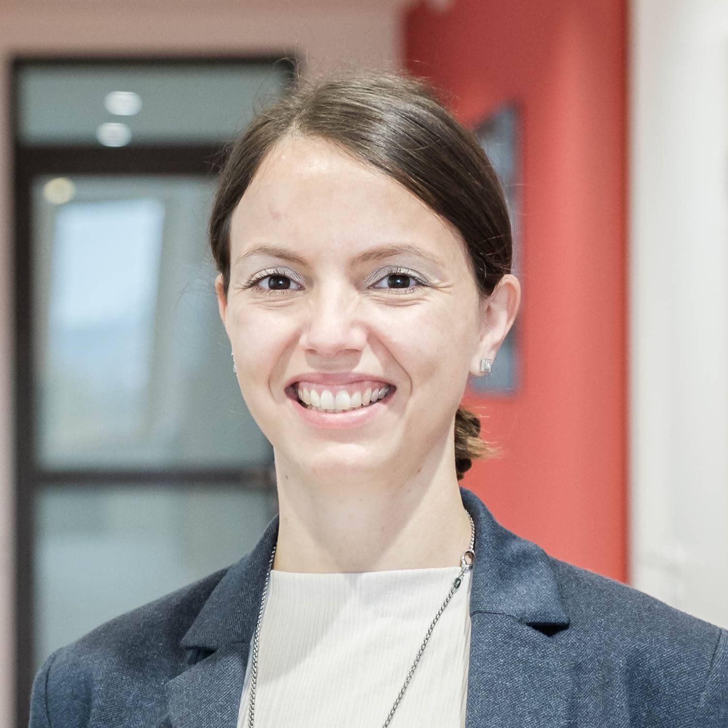 Eine freundlich lächelnde Frau mit heller Haut und zurückgebundenem braunen Haar steht in einem hellen Flur des modernen Medizincampus Passau. Die Frau trägt einen Blazer und ein helles Oberteil. Die Aufnahme vermittelt die offene und professionelle Atmosphäre des medizinischen Zentrums, das verschiedene Facharztpraxen, Diagnostikangebote und Gesundheitsdienstleistungen unter einem Dach vereint.